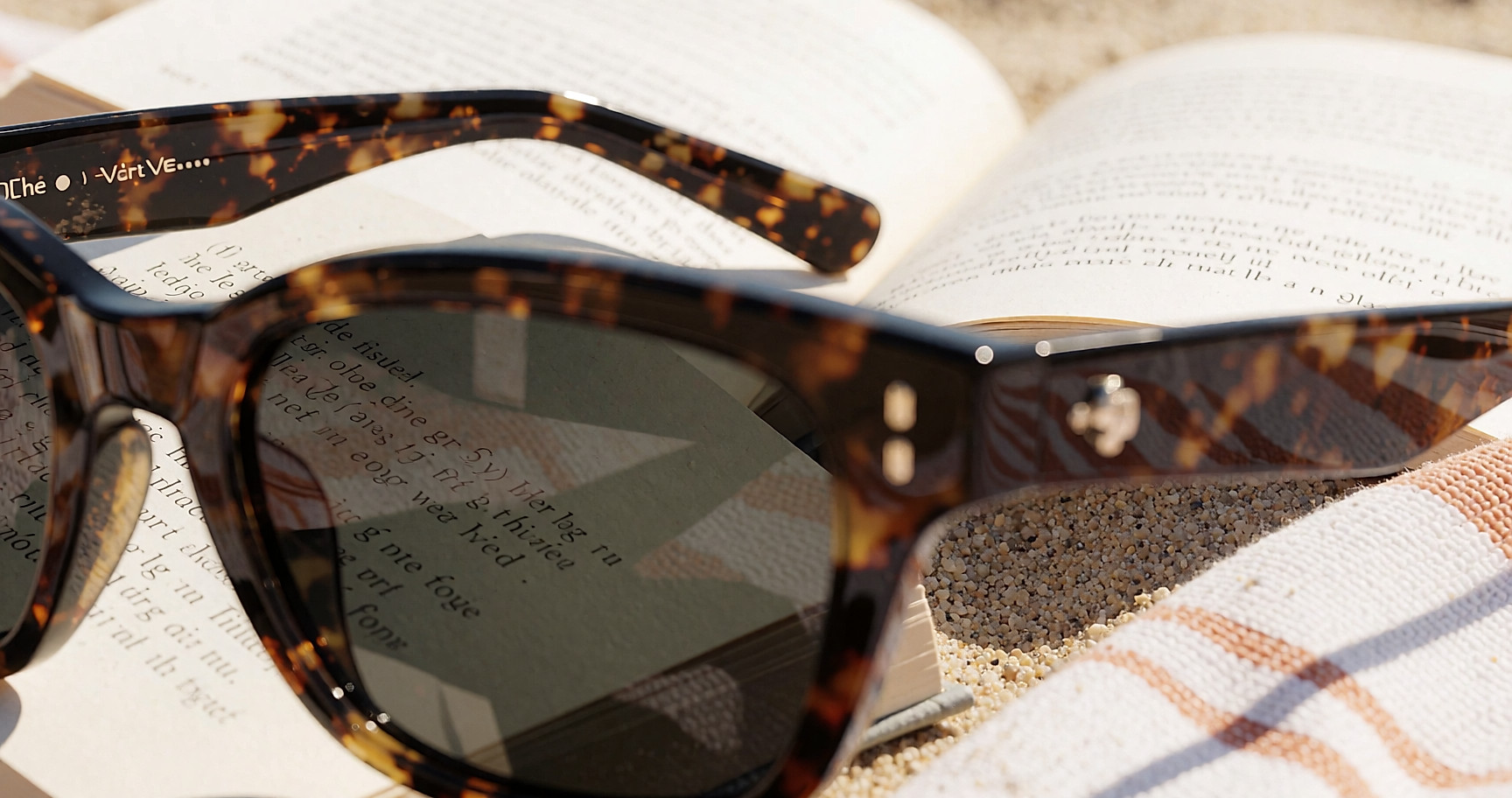 Lunettes de soleil à monture écaille posées sur un roman et une serviette, sur le sable