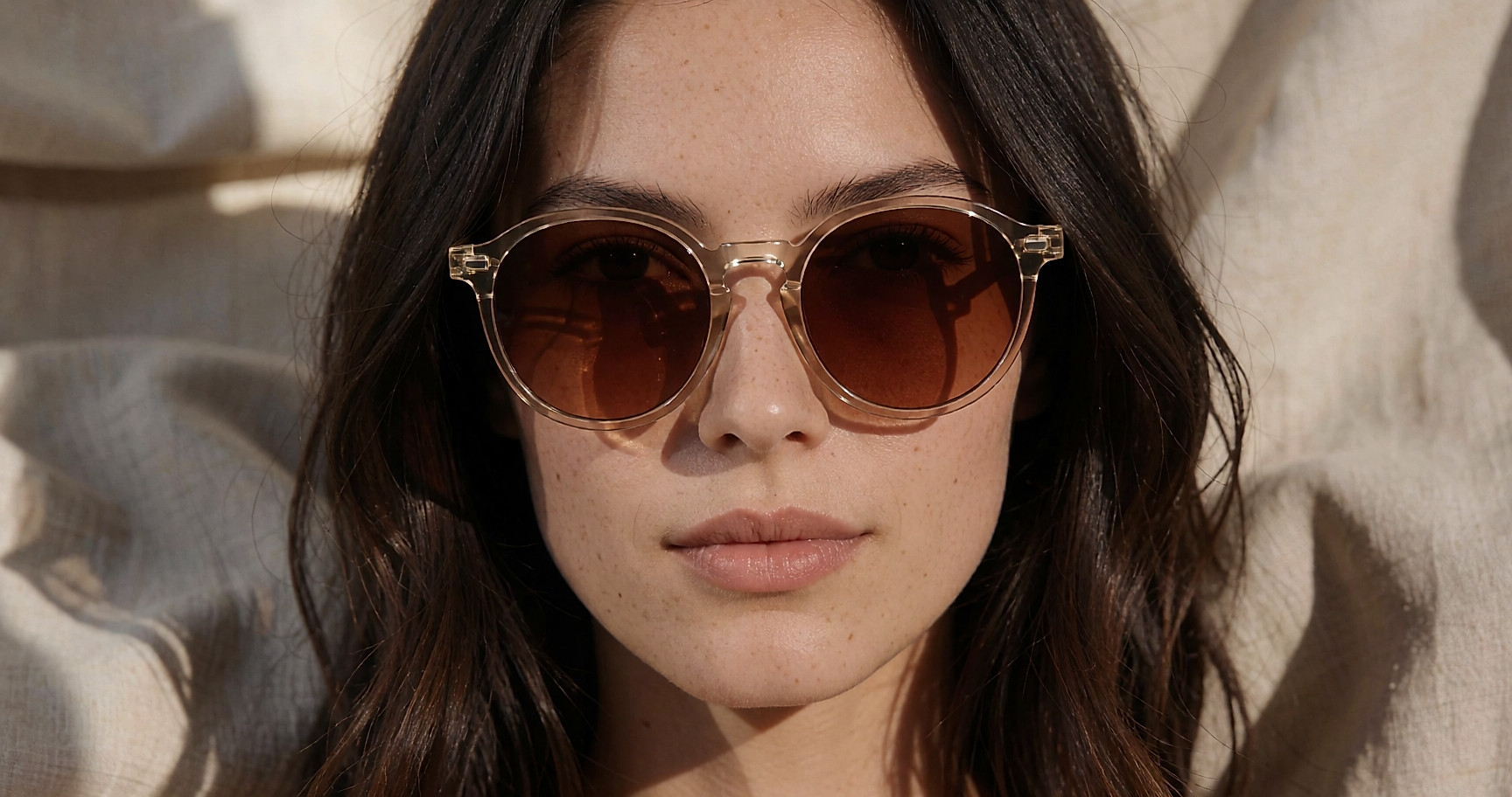 Lunettes de soleil rondes à monture fine translucide posées sur un lin beige froissé