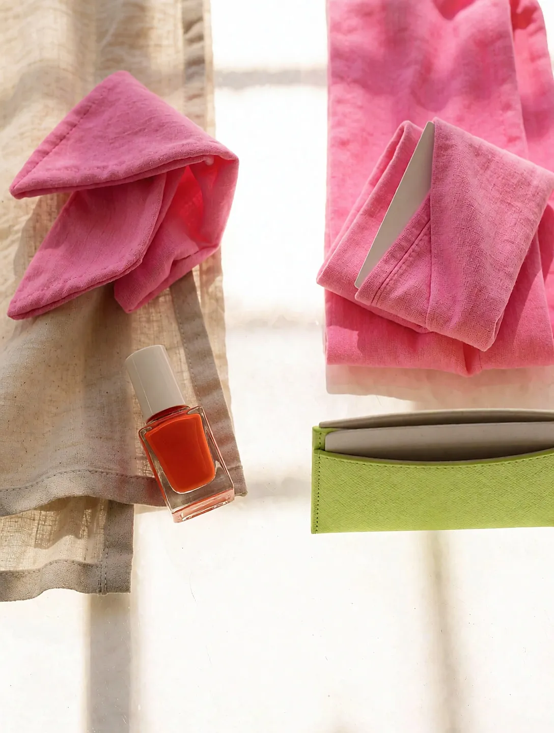Orange Nagellackfläschchen, rosa Haarreifen und neongrünes Kartenetui auf beigem Leinen