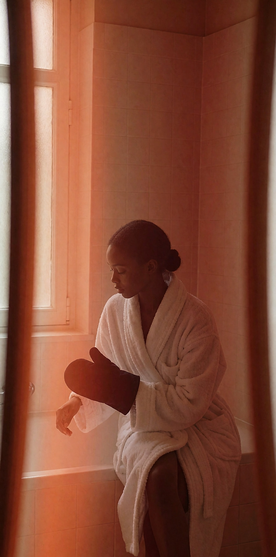 Femme en peignoir appliquant un lait autobronzant sur son mollet avec un gant en velours, dans une salle de bain claire