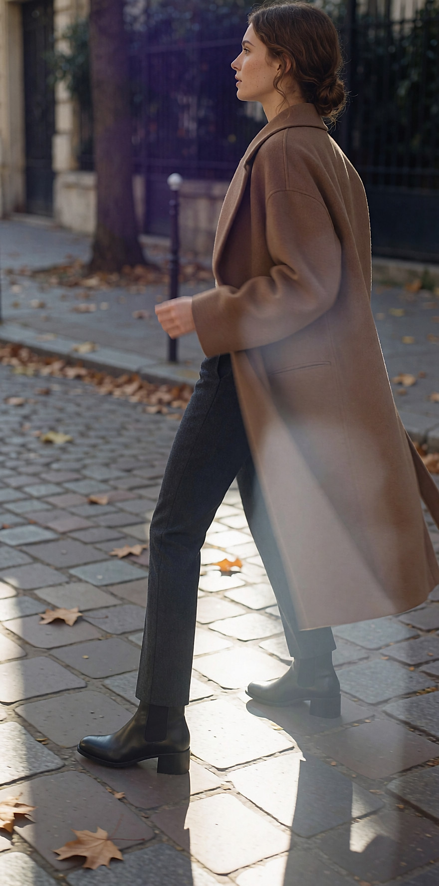 Jeune femme de dos sur un trottoir, pantalon slim gris, boots en cuir noir et manteau camel