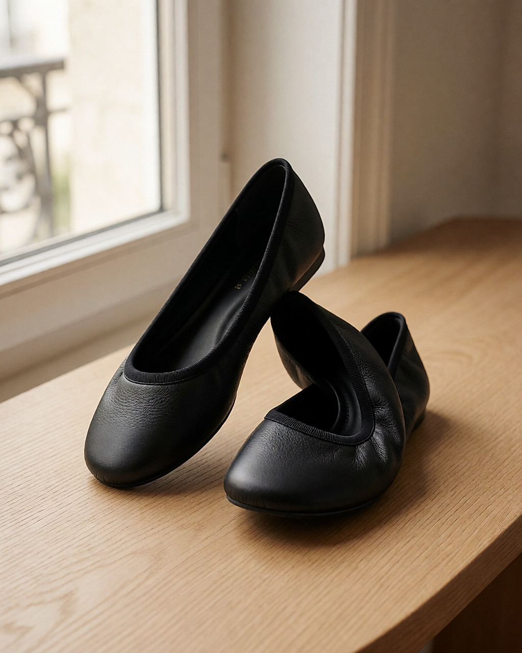 Deux ballerines en cuir noir souple sur une table en chêne clair, l'une pliée pour montrer la tige