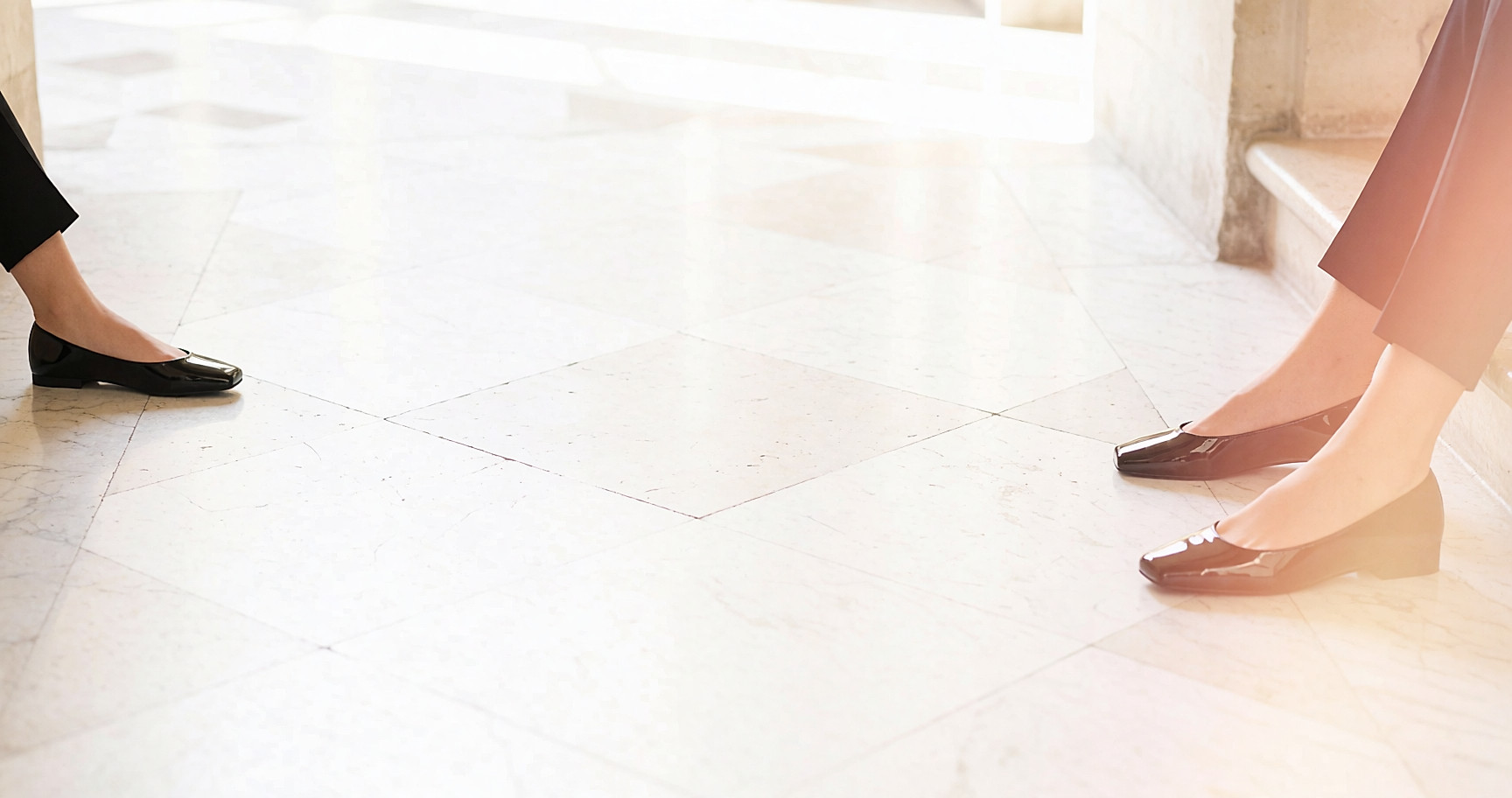Ballerines en vernis noir à semelle fine, jambes croisées dans un hall en marbre