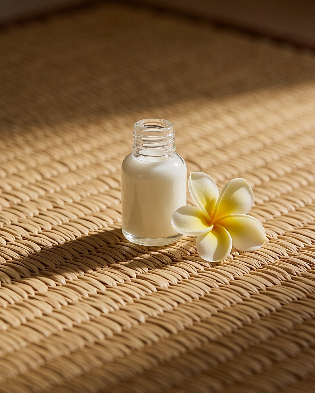 Mini flacon en verre de lait de monoï blanc avec une fleur de tiaré sur une natte