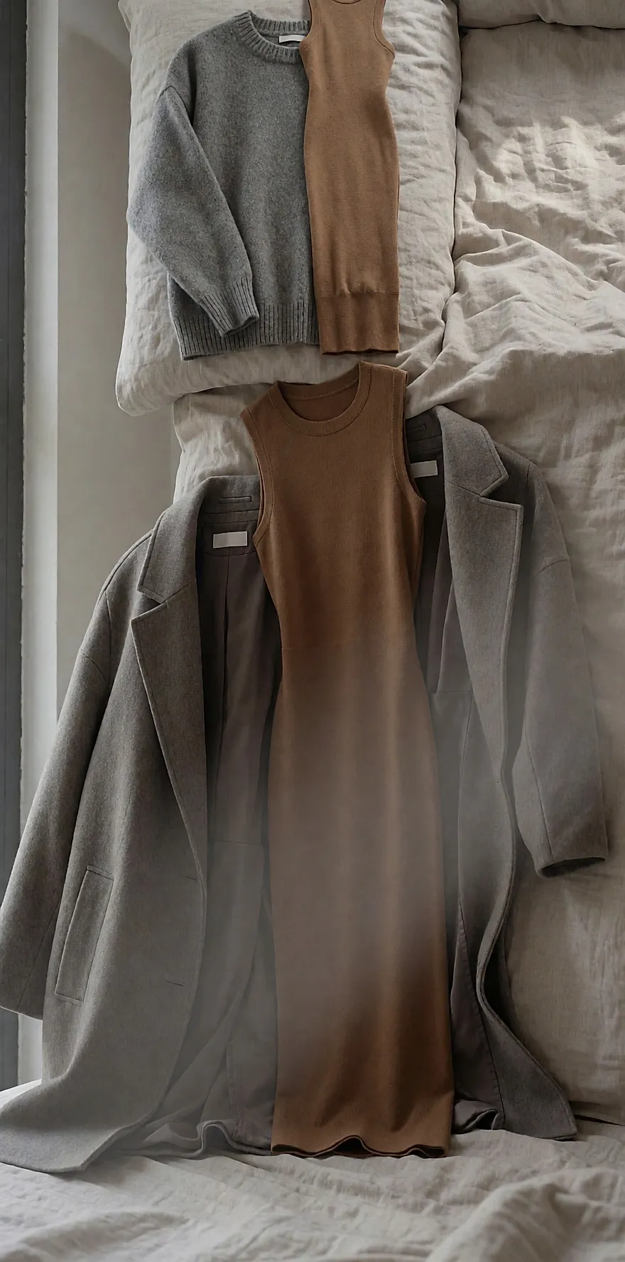 Grauer XXL-Pullover, eng anliegendes Strickkleid und strukturierter Mantel nebeneinander auf einem Leinenbett