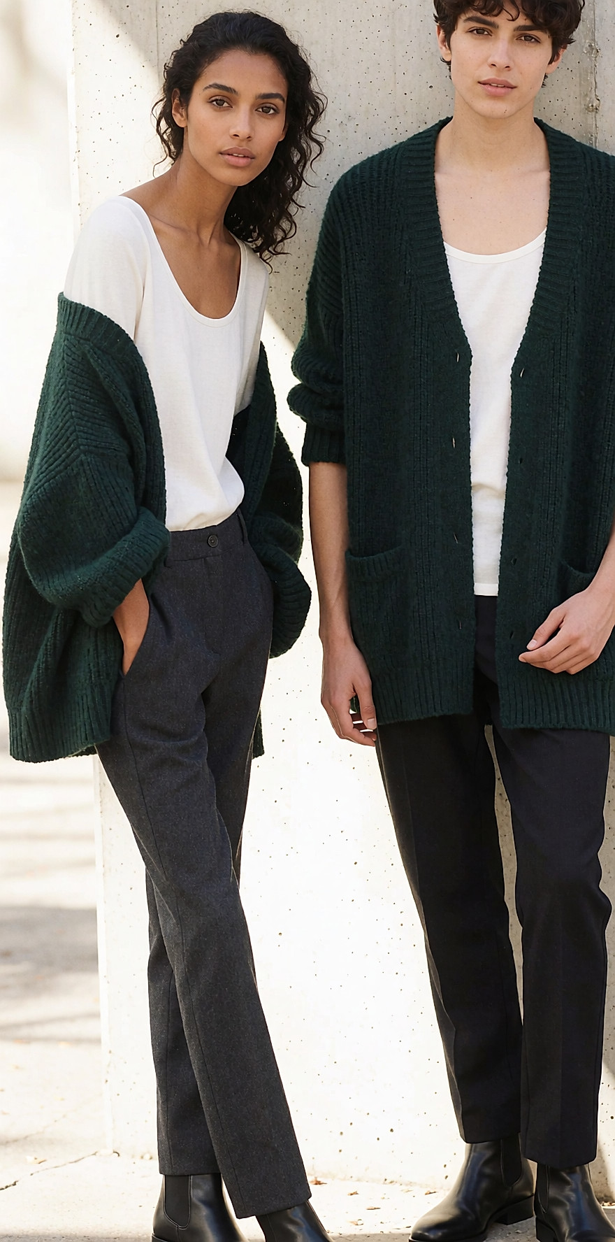 Silhouette en pull vert profond et pantalon anthracite adossée à un mur de béton