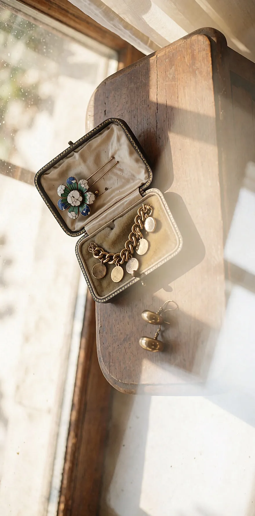 Boîte à bijoux ancienne ouverte sur une coiffeuse en bois, dévoilant une broche fleurie émaillée et un sautoir doré