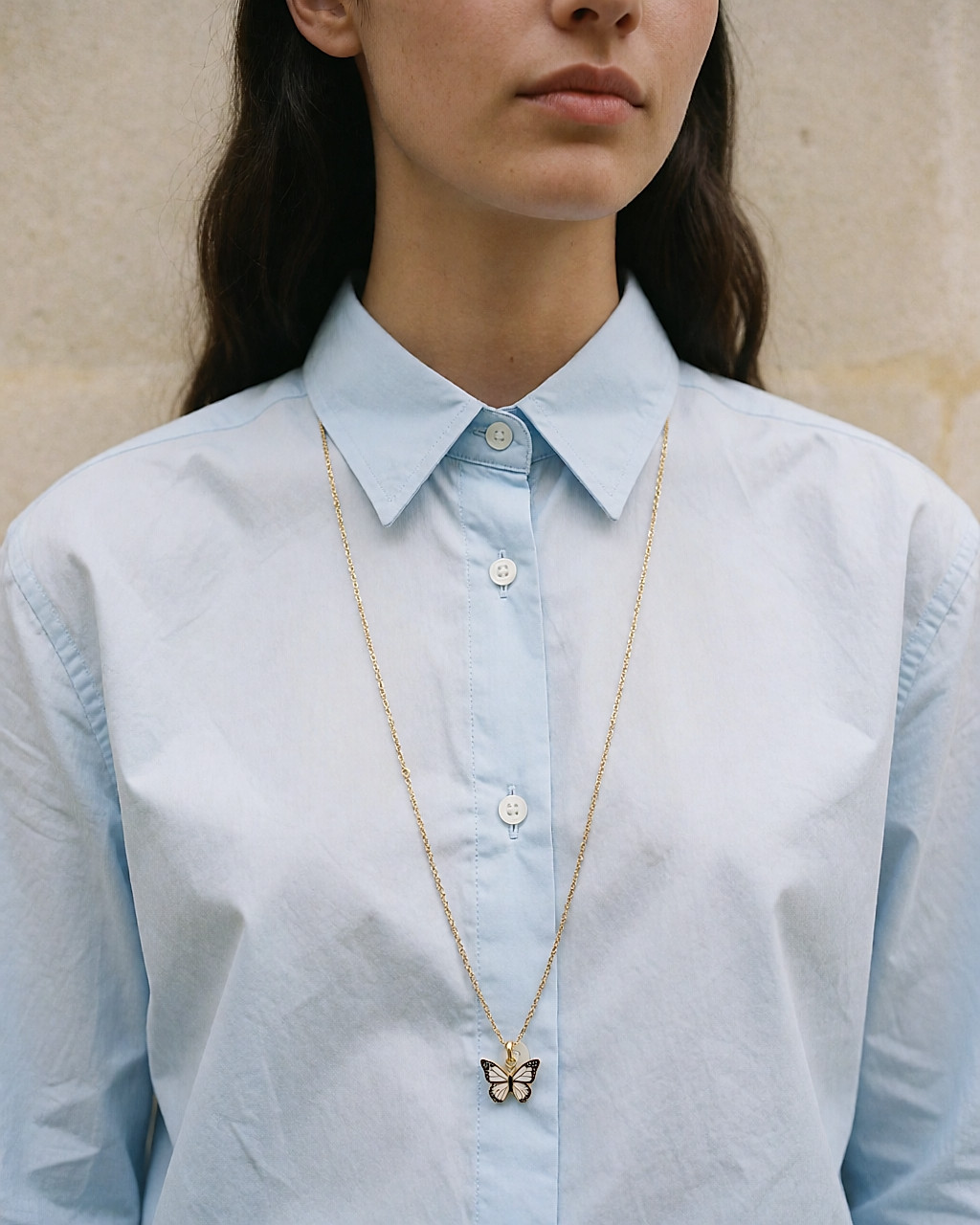 Sautoir doré à pendentif papillon émaillé porté sur une chemise en coton bleu pâle