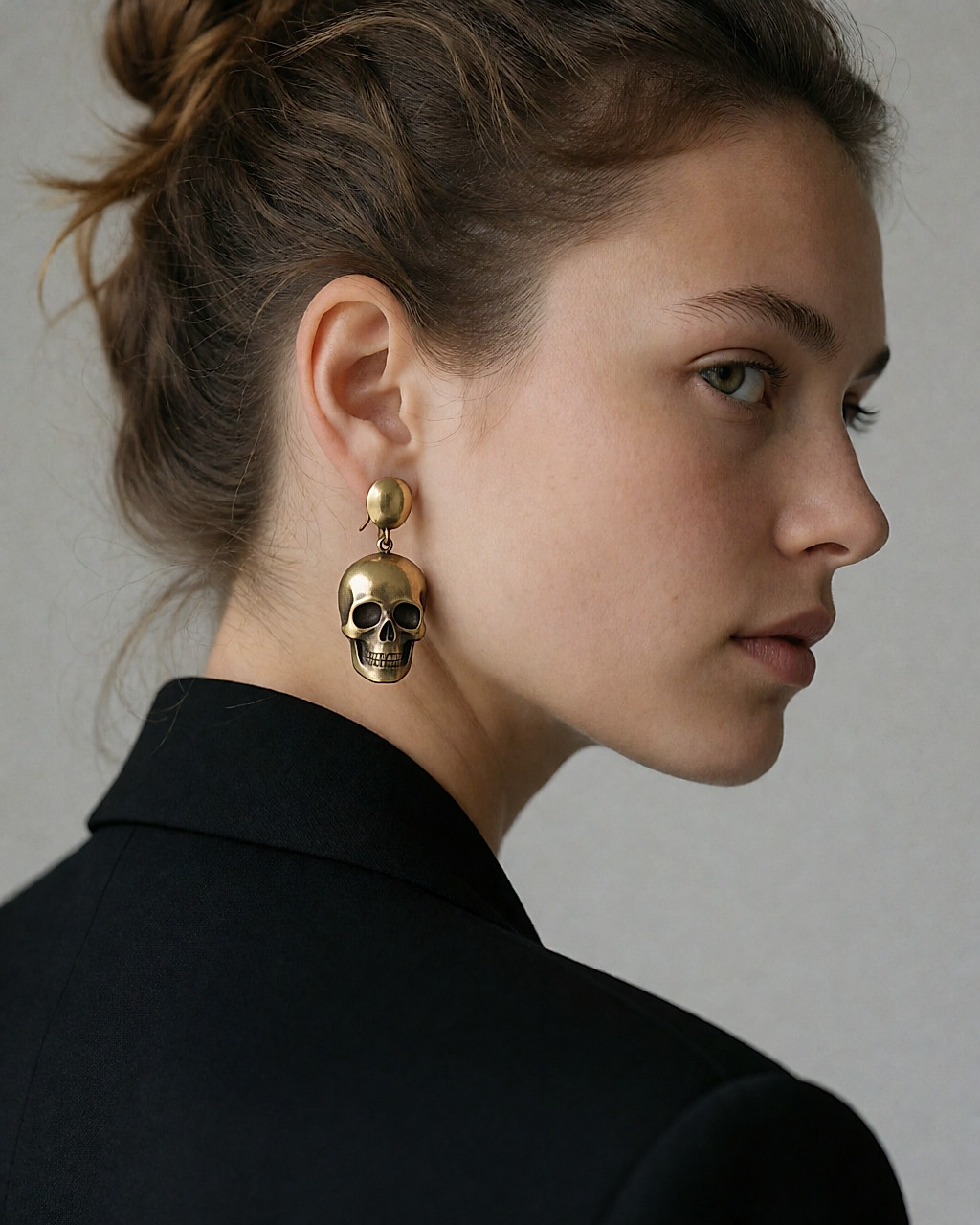 Boucles d'oreilles statement dorées à motif crâne, portées sur une oreille dégagée, cheveux relevés
