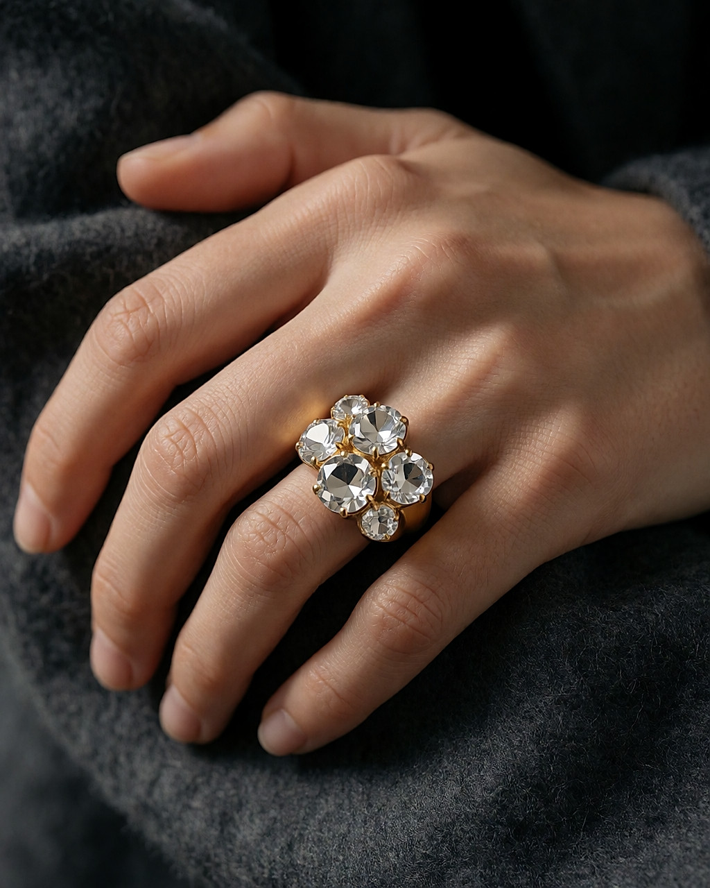 Bague statement dorée sertie de cristaux facettés, portée sur une main posée sur de la laine grise
