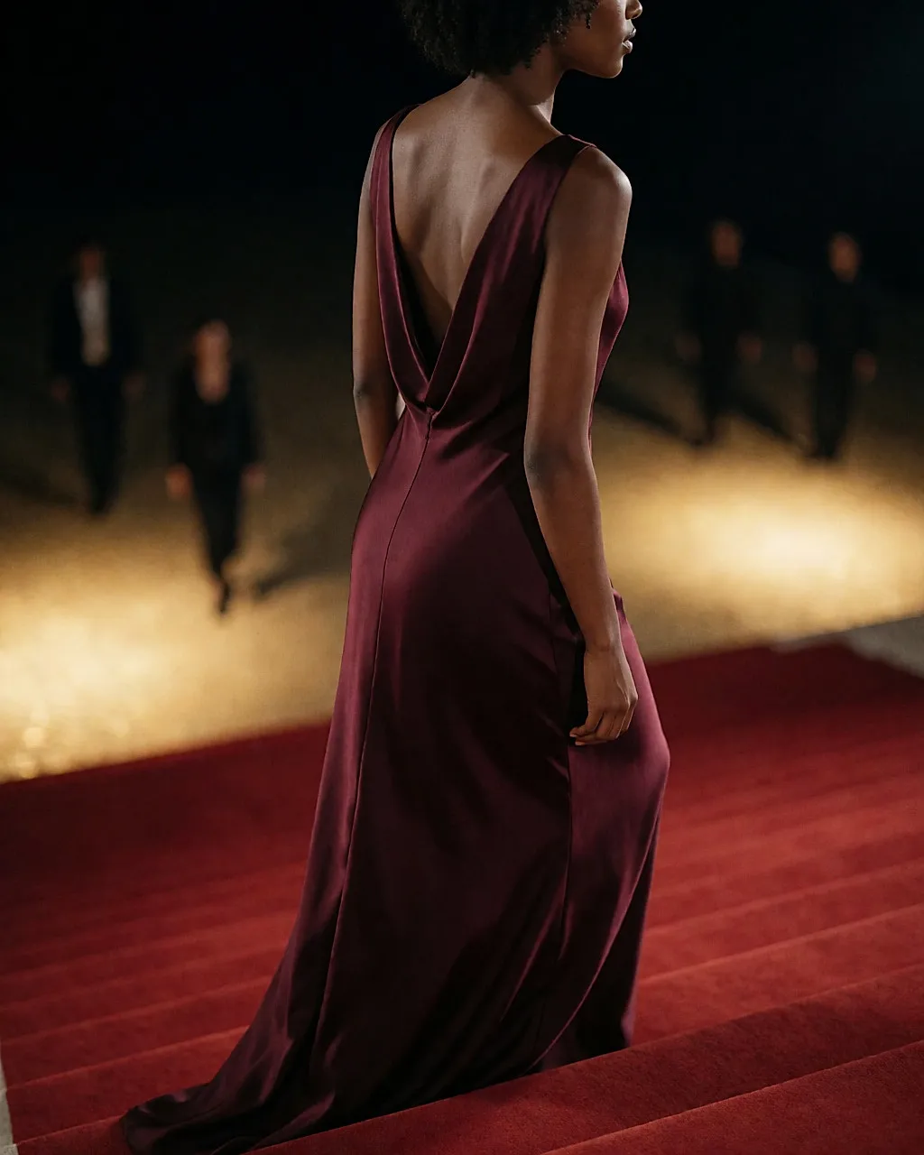 Rückansicht einer Frau in langem burgunderrotem Kleid, die auf einer Red-Carpet-Treppe im Scheinwerferlicht emporsteigt
