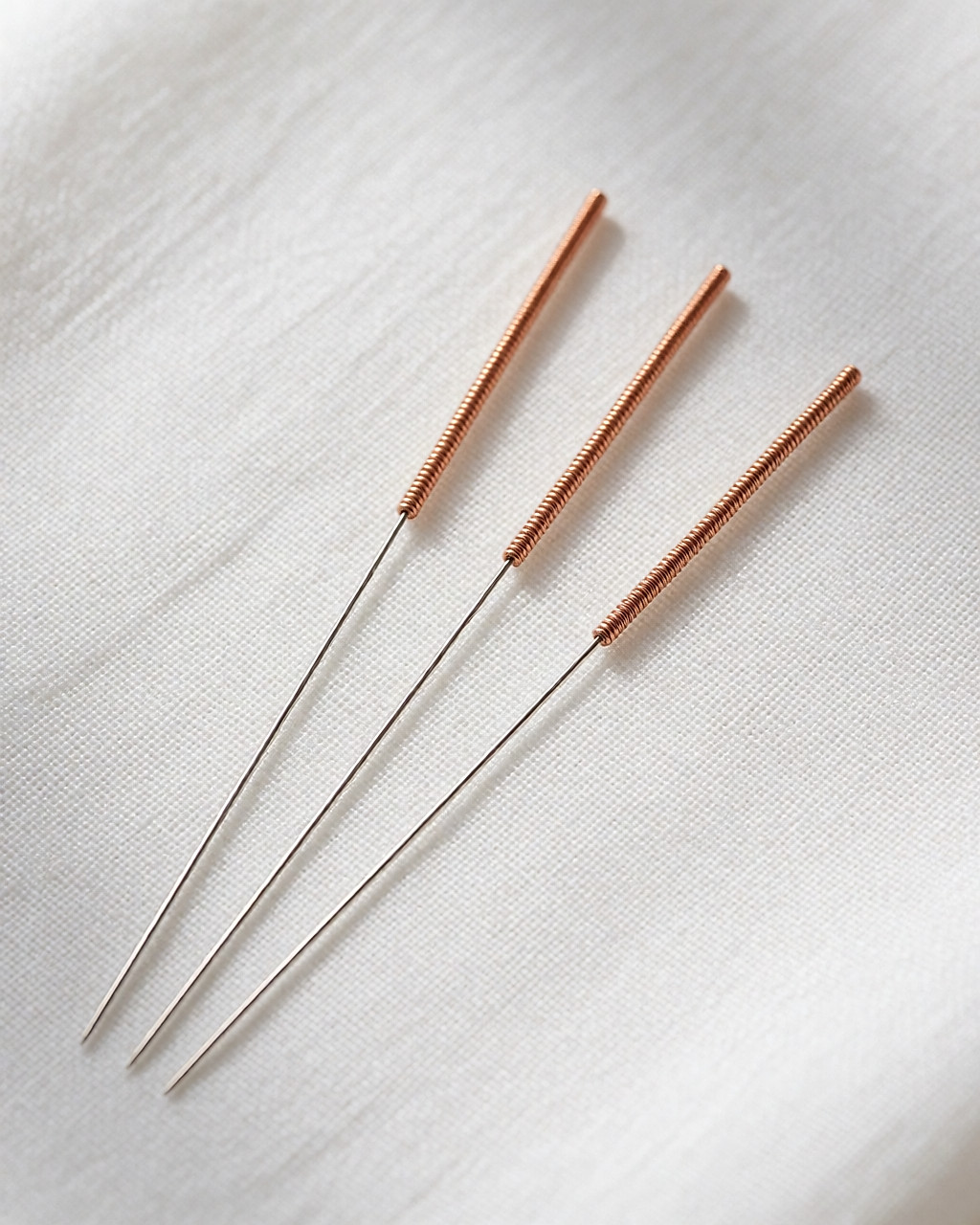 Trois longues aiguilles d'acupuncture très fines aux petits manches enroulés de cuivre, posées sur un linge de lin blanc