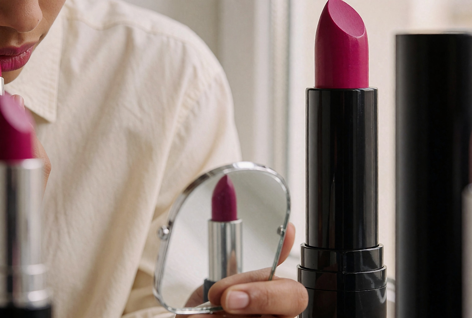 Jeune femme appliquant un rouge à lèvres fuchsia devant un miroir, près d'une fenêtre