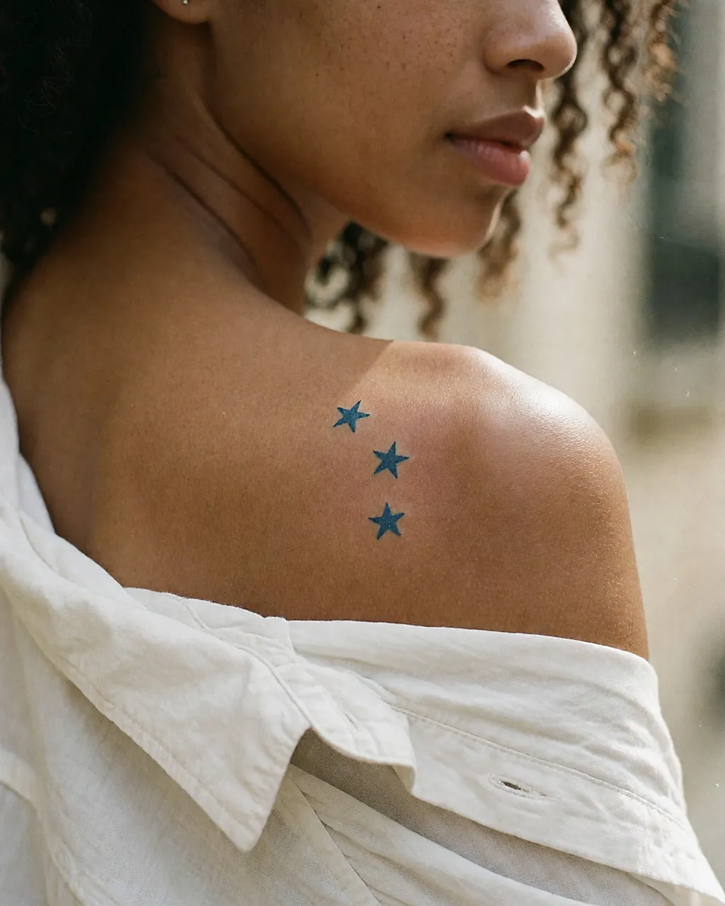 Tres estrellitas azules tatuadas en el hombro de una mujer joven, vista de tres cuartos por detrás