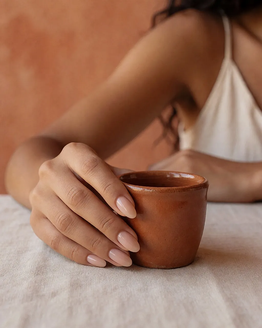 Manos con uñas en forma de almendra estilo babyboomer junto a una taza de barro, sobre fondo terracota
